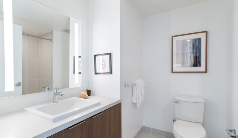 A compact apartment bathroom with a white sink on a wood vanity, a backlit mirror, a hand towel on a ring and a toilet beneath a framed print.