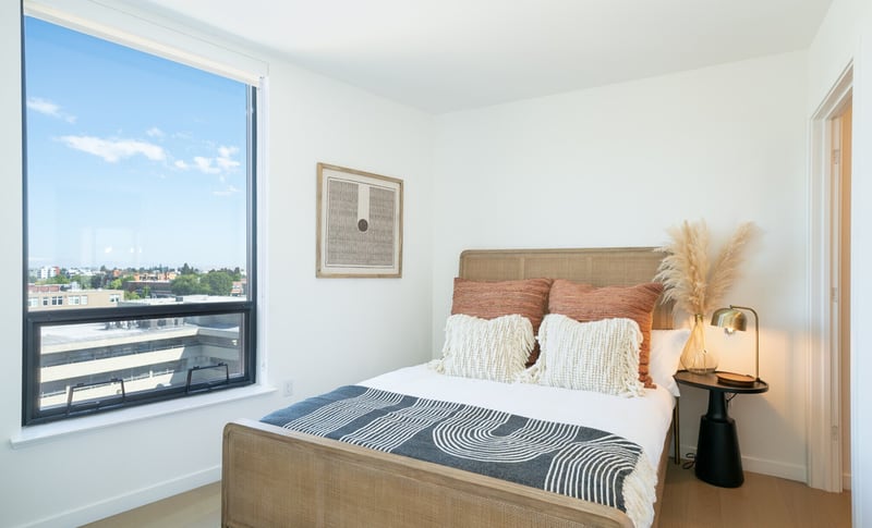 A staged bedroom in a new apartment, with a queen bed dressed in a patterned throw, a framed print on the wall and a large corner window looking out over a low-rise city skyline.
