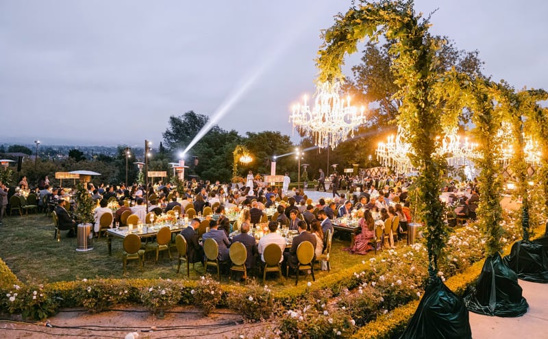 An outdoor evening wedding reception on a hillside lawn, with long banquet tables of seated guests set beneath crystal chandeliers hung from a leafy floral arch and strings of warm lights.