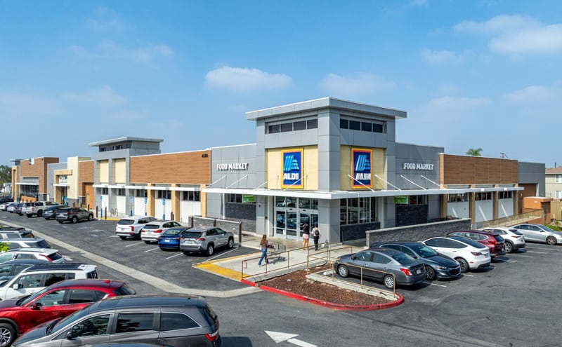 An Aldi food market with its blue logo on a grey and tan facade, shoppers pushing carts across a busy parking lot under a hazy sky.