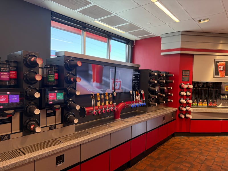 Roughly thirty linear feet given over entirely to self-serve dispensed beverage: bean-to-cup coffee towers with whole-bean hoppers, touchscreen grade selection, flavored latte taps on a manifold, and cup dispensers mounted at the point of dispense in three sizes. Every element removes labor from the transaction, which matters because hot dispensed runs about 62 percent gross margin, the top rung of the category ladder, and self-serve keeps that margin from being eaten by the labor line. Bean-to-cup also attacks the waste line directly, since nothing is brewed into a pot that gets discarded on a timer. Note the drainage trough running the full counter length: this is plumbed at construction, not retrofitted, and it is the single clearest reason a legacy 1,200 to 2,500 square foot box cannot copy this without being gutted. No Type I hood anywhere in the frame, which is the point. This entire profit center sits on the permissive side of the ventilation constraint that blocks most independents from hot food.